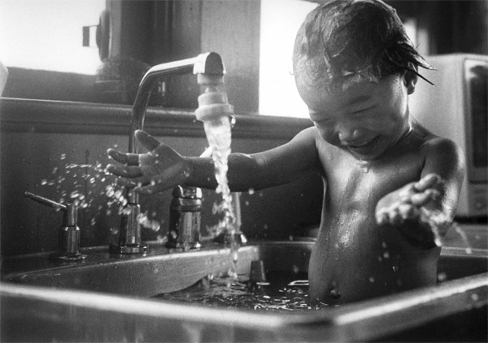 'In the Sink' alt='Black and white photograph of a naked baby playing joyfully while being bathed in a kitchen sink, taken by Laurie Rhodes'