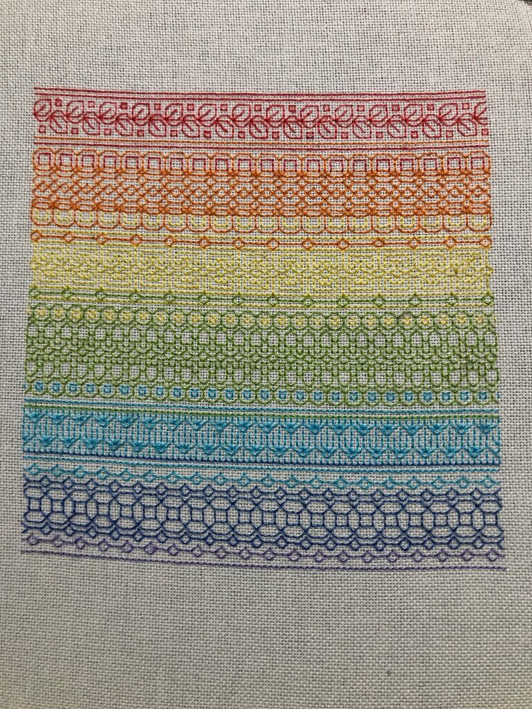 Seven bands of blackwork stitches in a rainbow of colors on white fabric