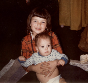 A young girl, about seven years old, holding her infant brother. She has white skin and shoulder-length brown hair and is wearing a red plaid top. The baby is white with brown hair and big, brown eyes. He's wearing a long-sleeved white top with spots of different blue hues.