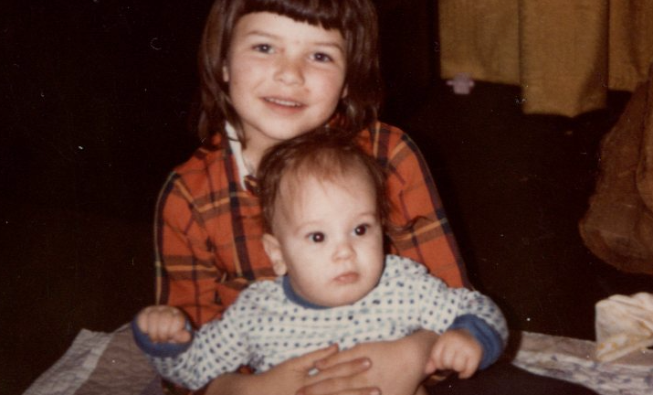 A young girl, about seven years old, holding her infant brother. She has white skin and shoulder-length brown hair and is wearing a red plaid top. The baby is white with brown hair and big, brown eyes. He's wearing a long-sleeved white top with spots of different blue hues.