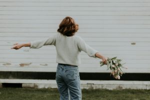 White woman shown from the back. She has dark blonde hair and is wearing a white sweater with faded blue jeans. She's holding a bouquet of flowers and has her arms flung out as if celebrating something.