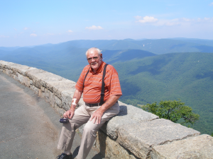 Image shows a tall, heavy-set white man, balding, with a fringe of white hair and a white mustache, wearing an orange shirt with tiny green and yellow stripes, khaki pants, and black suspenders. He's sitting on a low, stone wall with greenery and mountains in the background. 