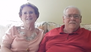 Image shows an older white couple smiling and sitting side by side on a loveseat. The woman has dark red hair and wears glasses. She's wearing a peach shirt with floral stitching around the collar and a long, gold chain with a round locket. The man  has a receding hairline with white hair and mustache. He wears glasses and a red, collared shirt. 