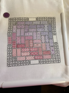 White fabric in a rectangular plastic frame (Q-Snap) with a black outline mostly filled in with rectangles of blackwork in varying colors.